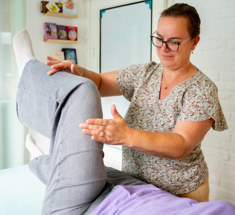 Een vrouw biedt een therapeutische behandeling aan een liggende patiënt in een lichte kamer. Kinesiologiebehandeling
