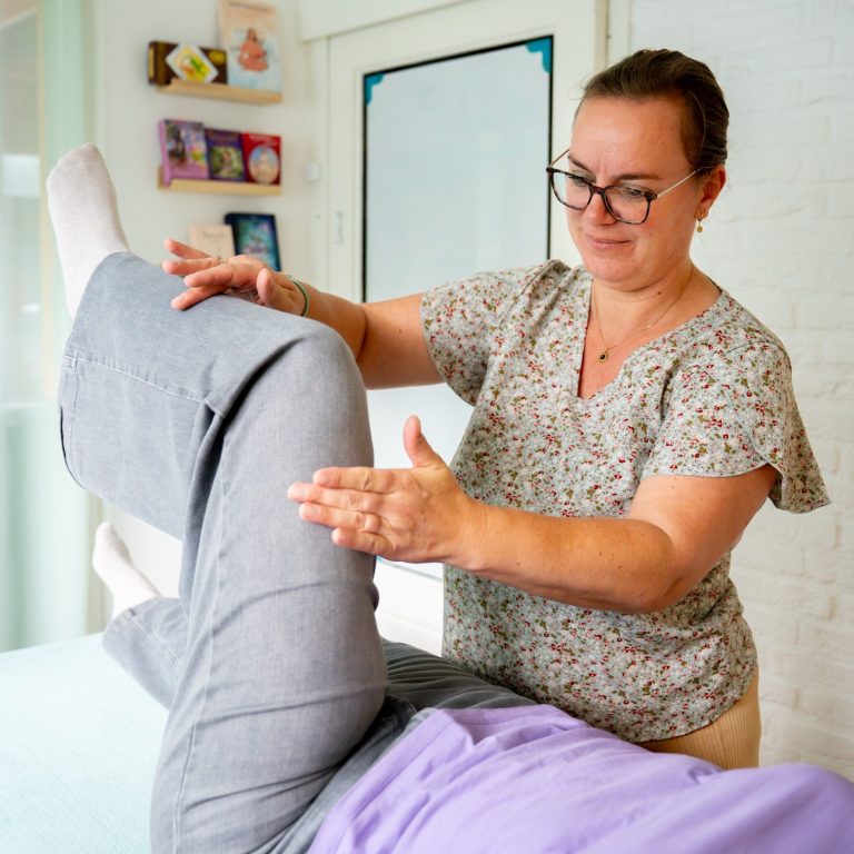 Een vrouw biedt een therapeutische behandeling aan een liggende patiënt in een lichte kamer. Kinesiologiebehandeling
